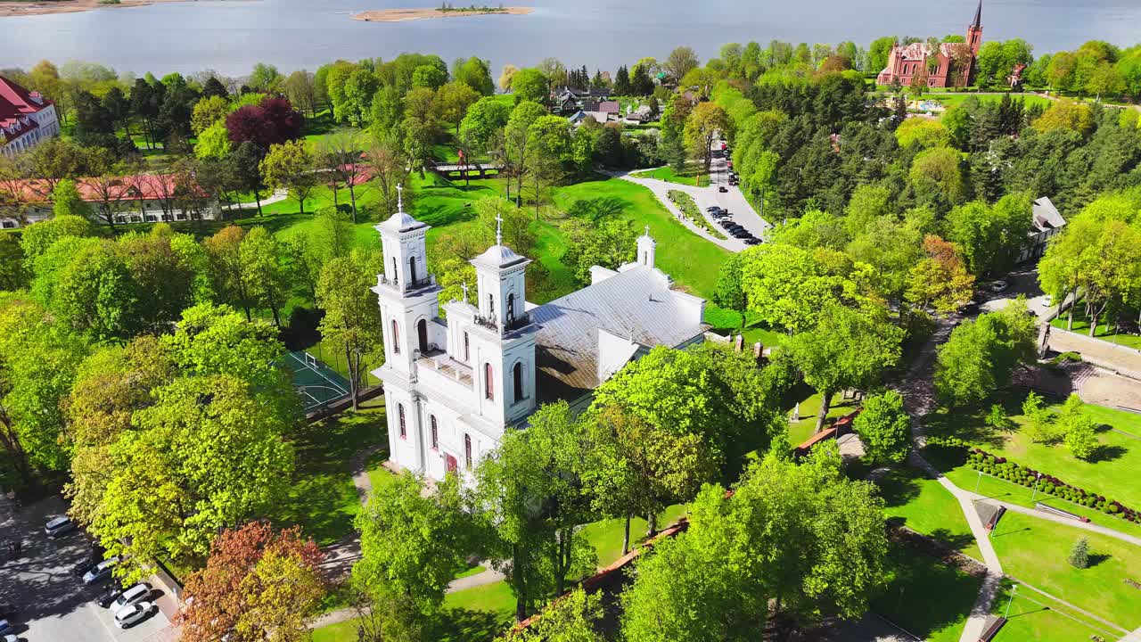A bright white church with twin towers sits at the heart of a green park near a riverside, framed by spring trees, walkways, and nearby town buildings.