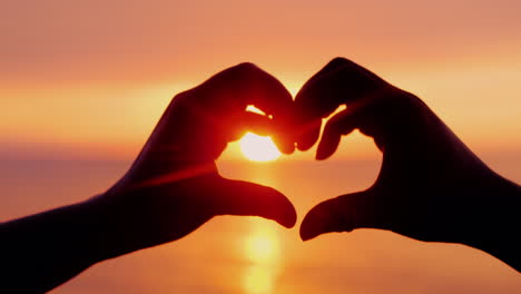 two hands show the shape of the heart around the sun setting over the sea