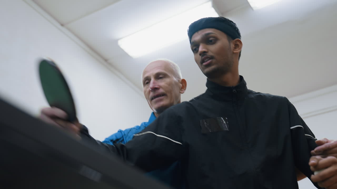 Elderly coach guiding young player on proper racket control during tennis training indoors, focusing on grip and precision with determination in their expressions under bright indoor lighting