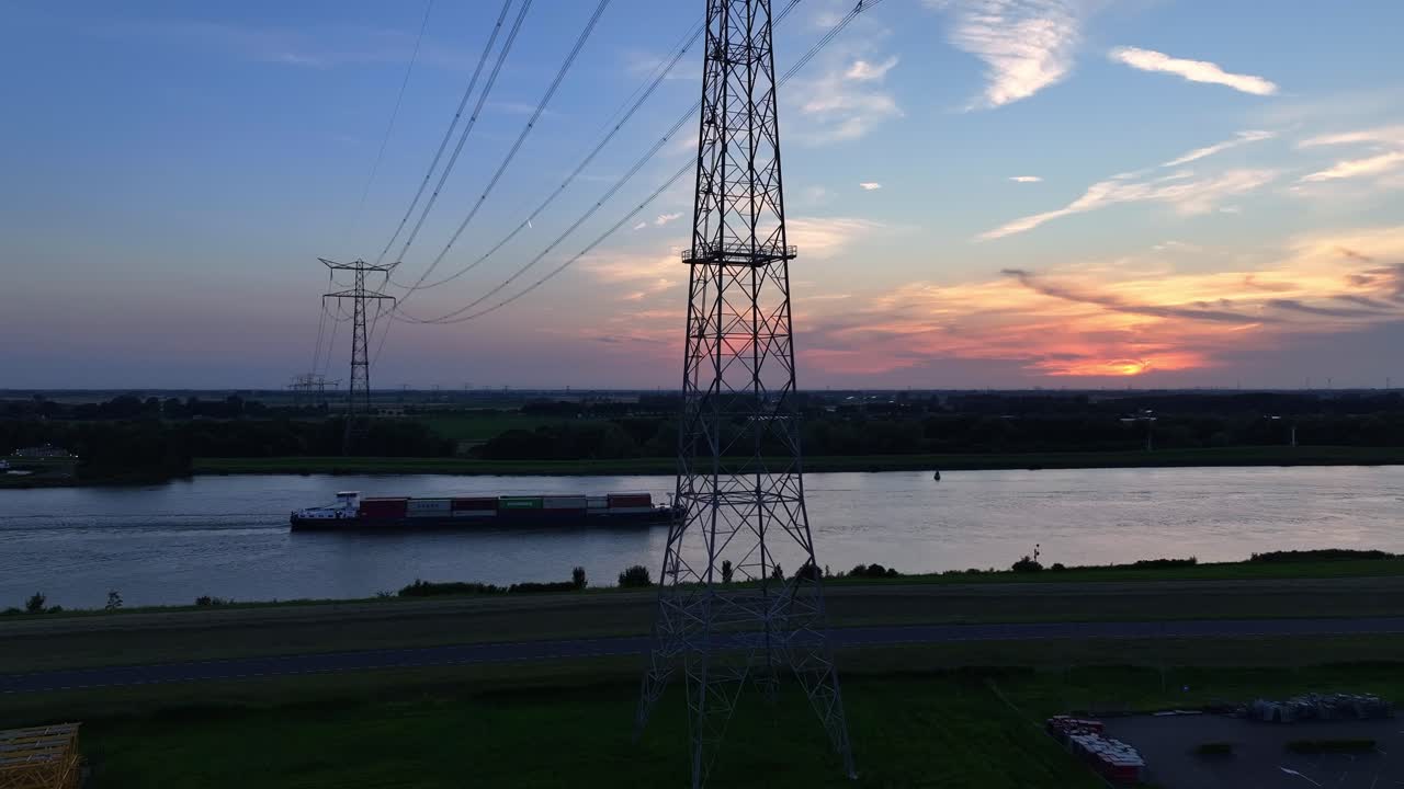 A barge moves along a calm river at sunset with power lines crossing overhead