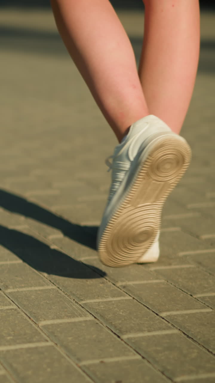 Back view of individual wearing white canvas shoes walking on interlocked path with shadow cast on the ground, sunlight illuminates legs and pathway