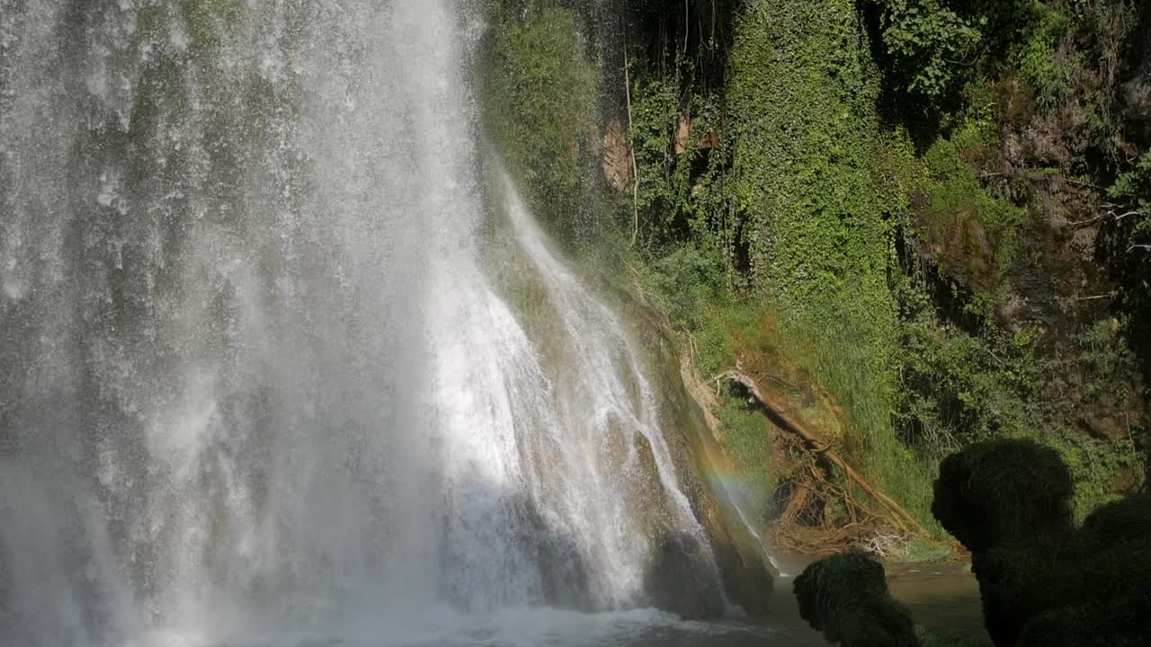 Beautiful downpour cataract waterfall on plunge pool. Water stream drop on rocks and pool surrounded by vegetation. Sun rays create a rainbow colour spectrum halo on one side