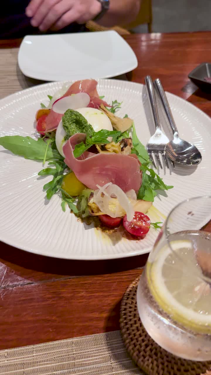Ripresa dall'alto di una mano che sistema le posate accanto a un piatto di insalata gourmet con burrata