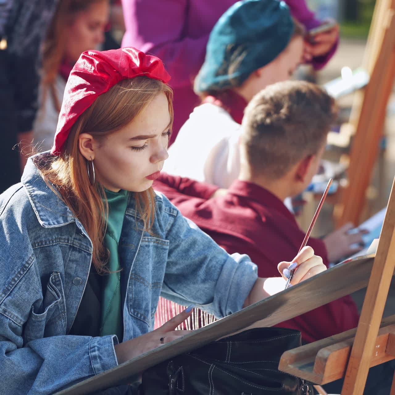 Young artists paint on easels in the city. Pretty woman artist is painting picture with brush and watercolors in the busy street. Side view of female artist.