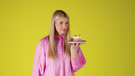 young blonde woman smelling a piece of delicious cake on plate, colorful studio portrait