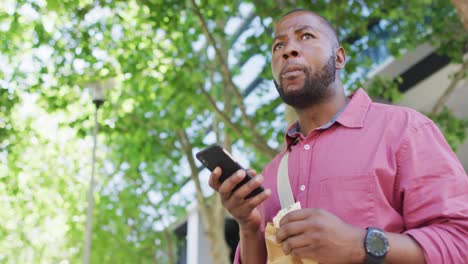 Hombre-Afroamericano-Comiendo-Y-Usando-Un-Teléfono-Inteligente-En-La-Ciudad