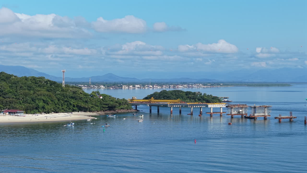 Ponte de Guaratuba bridge under construction, spanning Guaratuba Bay in Paraná, Brazil, with forested shoreline, calm waters, blue sky, and boats in the distance, wide drone shot