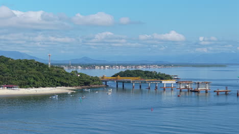 ponte de guaratuba bridge under construction, spanning guaratuba bay in paraná, brazil, with forested shoreline, calm waters, blue sky, and boats in the distance, wide drone shot