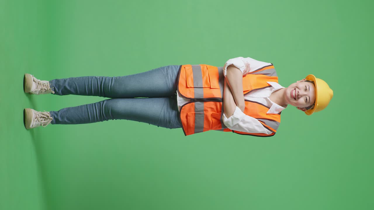 cuerpo lleno de ingeniera asiática con casco de seguridad cruzando los brazos y sonriendo a la cámara mientras está de pie en el estudio de fondo de la pantalla verde