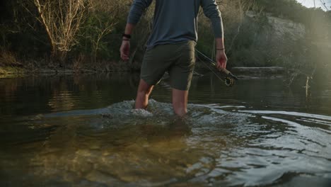 male fly fisherman steps into sun soaked lake water with sandals close up slow motion