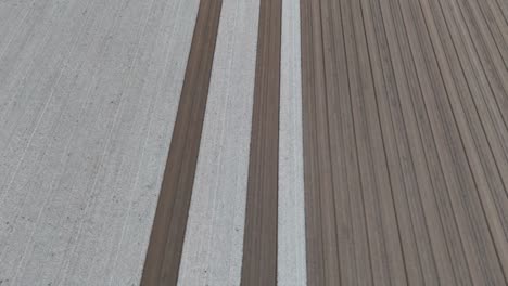 slow-motion drone shot reveals perfectly parallel strips of pale tilth and rich brown earth freshly leveled across a vast field forming an orderly grid poised for the next planting season
