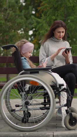 woman checks email while little girl in wheelchair watching videos on smartphone. focused mother and daughter use gadgets after walk in city park