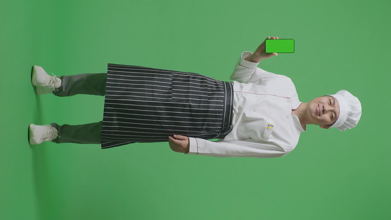 cuerpo lleno de hombre asiático chef sonriendo y mostrando pantalla verde teléfono inteligente a la cámara mientras estaba de pie en el estudio de fondo pantalla verde