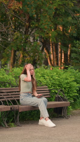 mujer sentada en un banco de madera al aire libre, sosteniendo el teléfono en su mano derecha mientras ajusta su cabello con su izquierda, rodeada de exuberante vegetación y árboles, con la construcción