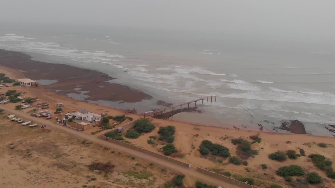 Forward angled drone approach arriving at the shore of Allana Gadoor Village Beach (Teen Khaji), featuring a long simple pier extending into the ocean with waves breaking against a hazy horizon