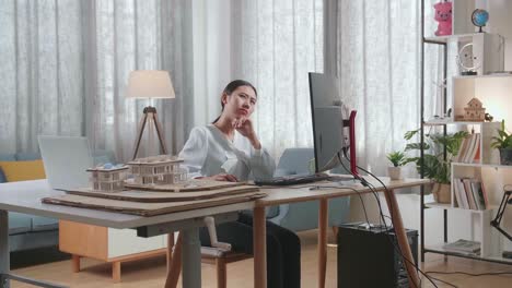 asian woman engineer with the house model thinking while working on a desktop at home