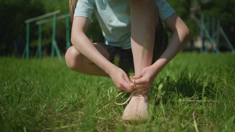 close-up view of a little girl tying her shoelace on a grassy field, with a blurred background of trees and playground equipment