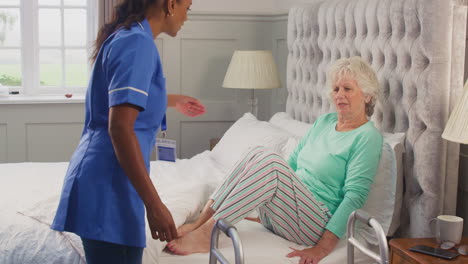 senior woman at home using walking frame being helped out of bed by female care worker in uniform