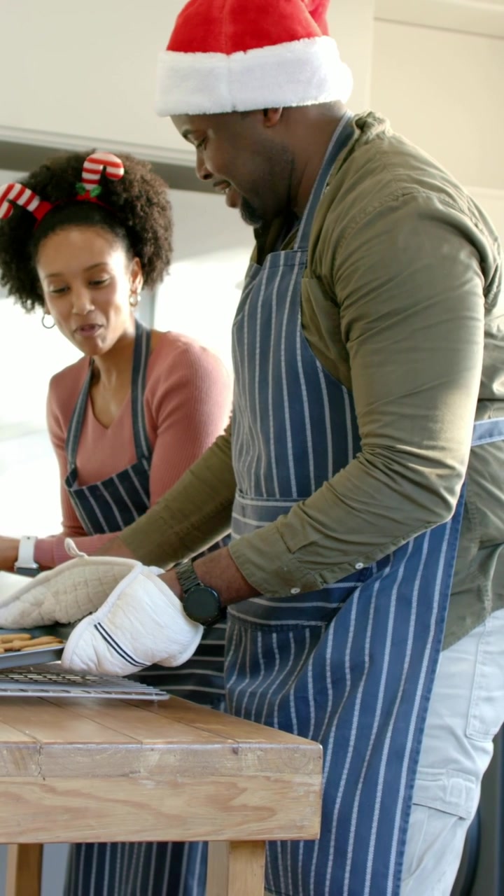 Video verticale: Uomo con cappello di Babbo Natale che toglie una teglia dal forno in cucina, posandola sul tavolo mentre una donna controlla.