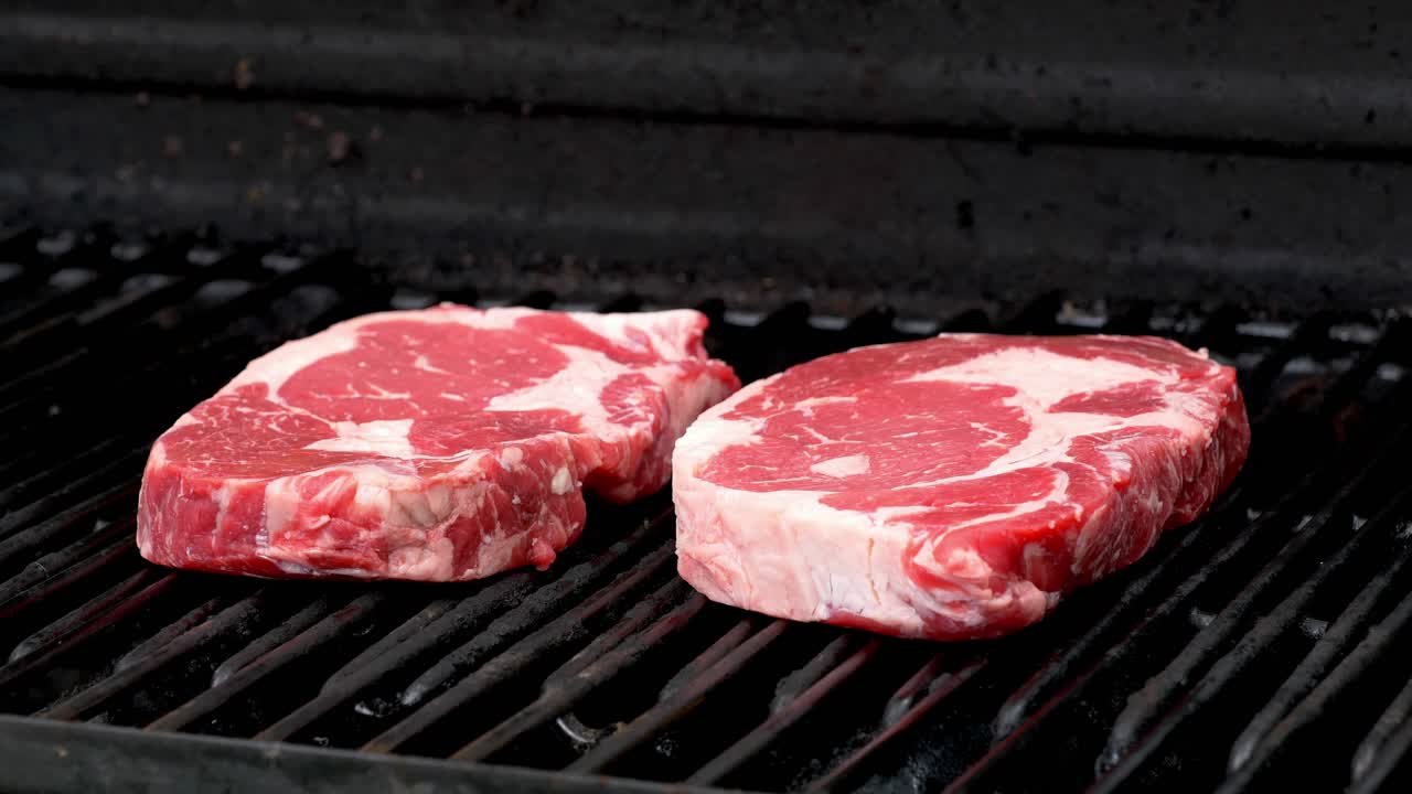 Two raw rib-eye steaks getting ready to cook on a grill