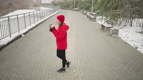 lady in red hoodie opens water bottle, drinks from pink bottle outdoors in serene winter park setting with snow-covered ground, iron railing, benches, and trees in peaceful misty atmosphere