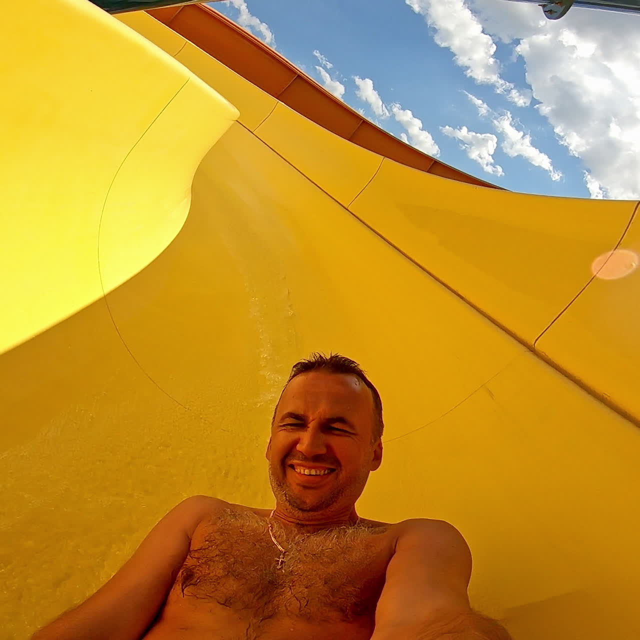 Joyful male going down from yellow pipe outdoors. Man having fun and sliding down in a water slide in summer at a sunny day.