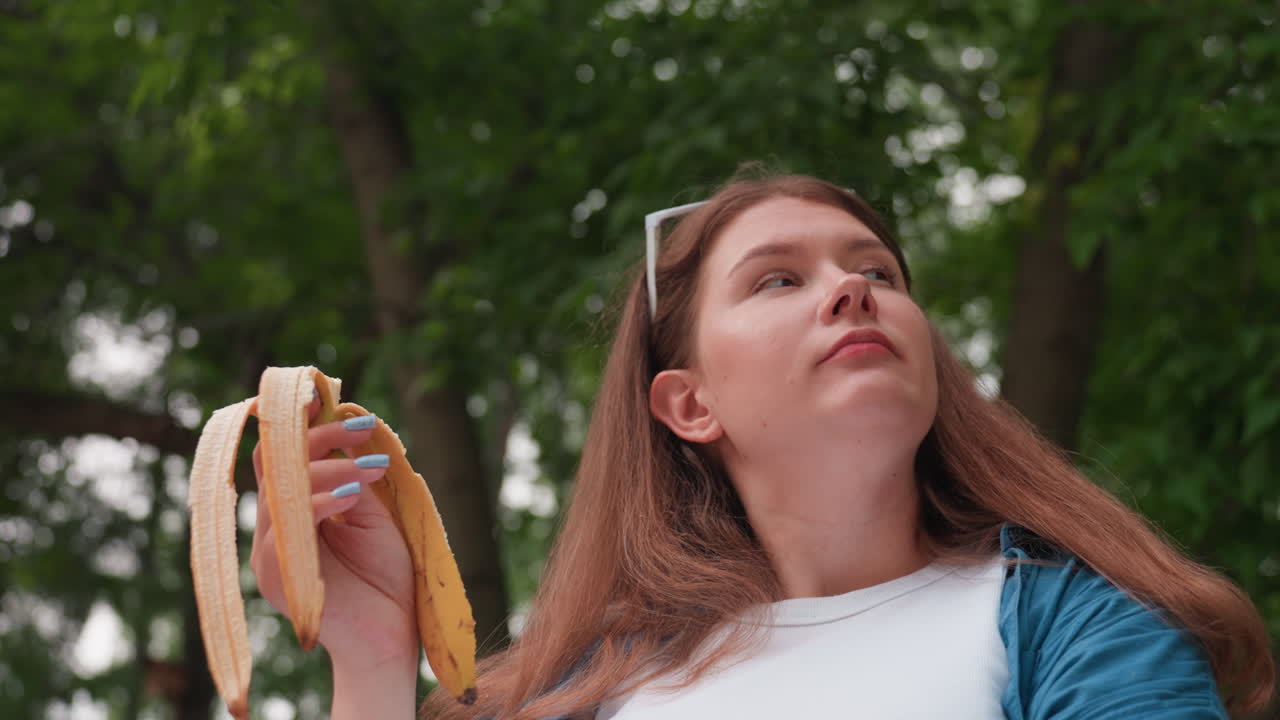 푸르른 녹음이 우거진 나무 아래에서 차분하고 사려 깊은 표정으로 주변을 둘러보며 플랜틴을 먹고 있는 여성, 평화로운 사색의 분위기를 고조시키는 자연광