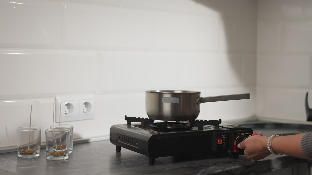 Close up of female hand holding stainless steel pot above gas burner while preparing to cook, carefully checking if gas flame is lit in well-lit modern kitchen with utensils and candles on counter