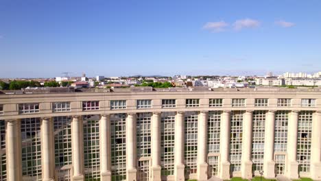 A-drone-camera-flies-over-a-square-and-an-architecturally-magnificent-building-on-the-Place-de-l'Europe-in-Montpellier,-France,-revealing-the-city-in-the-background