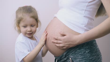 Glückliches-Mädchen-Spielt-Mit-Dem-Bauch-Der-Schwangeren-Mutter,-Der-An-Der-Wand-Steht