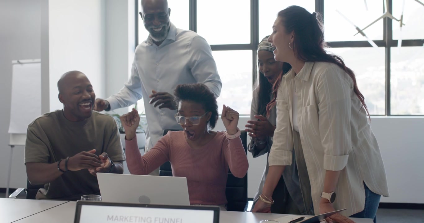 Diverse team in business attire cheering around woman operating laptop at table after funnel win