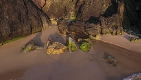 Water-running-into-sea-from-cliffs-along-sandy-beach-meeting-the-breaking-waves-at-evening-sunset-aerial-pull-back-shot