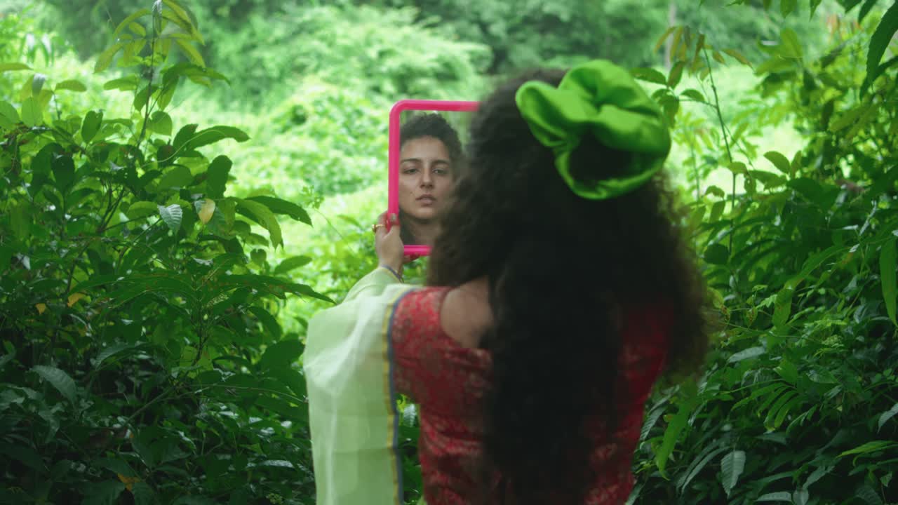 Camera moves closer to a woman in sari with loose hair and green accessory, seen from behind, holding a mirror reflecting her face amid dense vegetation