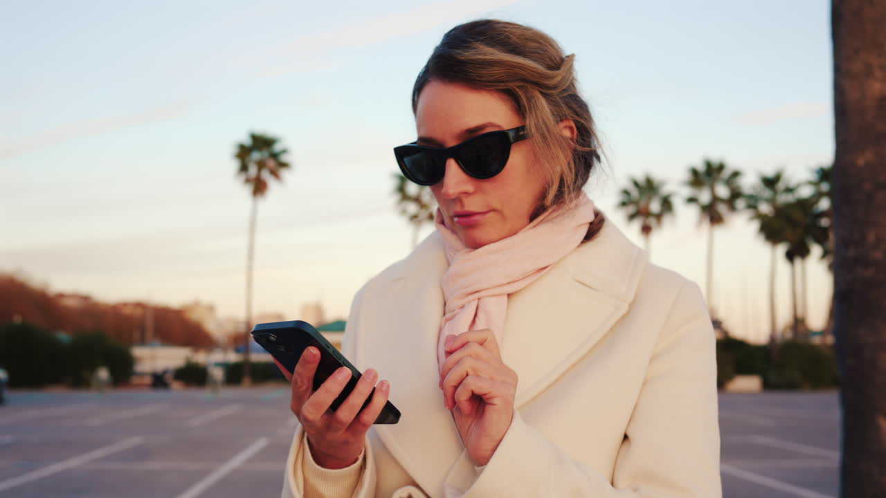Donna elegante con cappotto bianco, sciarpa e occhiali da sole che controlla il telefono in una zona esterna soleggiata con palme