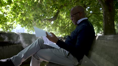 Bald-Black-Man-relaxing-on-a-bench-during-a-lunch-break,-vaping-and-reading-under-bright-green-trees-in-a-park