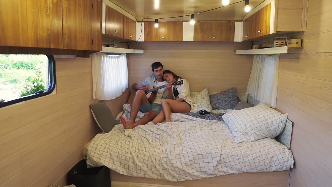 Slow motion of young just married couple wearing casual having fun to playing guitar in the bedroom in the wheels house. Girl sensually listen to her husband, leans on him