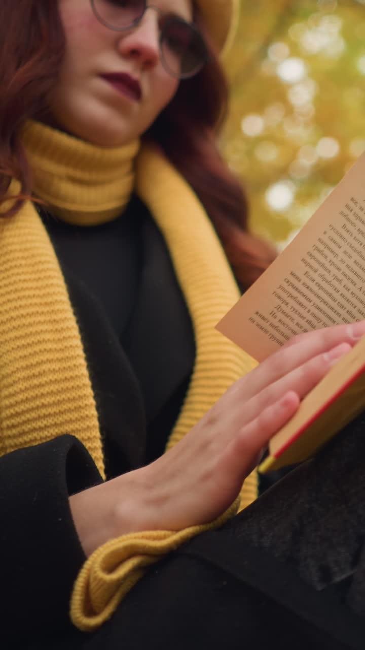 primer plano de una mujer con gafas de sol y una bufanda cálida, leyendo pensativamente un libro cubierto de amarillo, su mano descansa suavemente en las páginas mientras el fondo presenta árboles suavemente borrosos