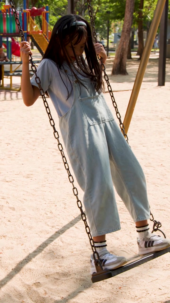 Una ragazza asiatica allegra in salopette di jeans sta in piedi su un'altalena al Parco Lumpini, Bangkok.