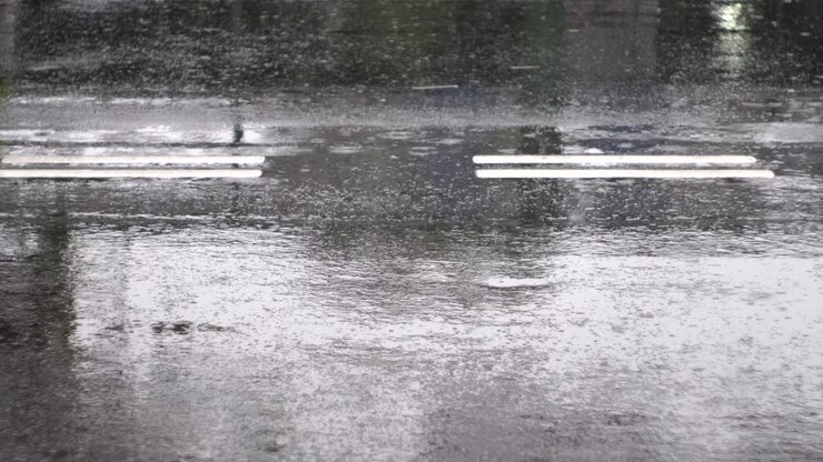 Raindrops causing ripples, falling on reflective black tarmac road with white road markers