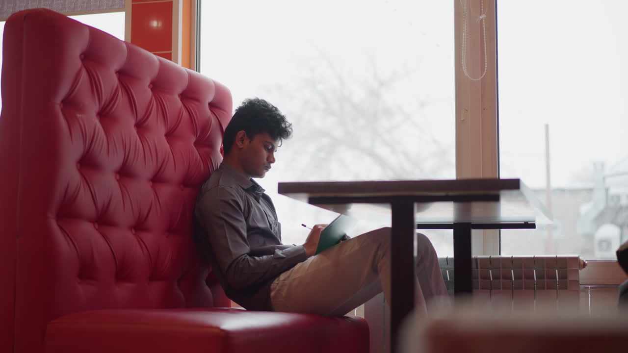 Student sitting on red cushioned bench by window, writing in notebook with pen, snow visible outside, calm indoor setting suggests focus, reflection, study moment in relaxed environment with natural light