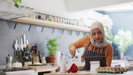 Video-Einer-Glücklichen-Gemischtrassigen-Frau-Im-Hijab,-Die-Zu-Hause-In-Der-Küche-Backt-Und-Dabei-Ein-Rezept-Auf-Einem-Tablet-Verwendet