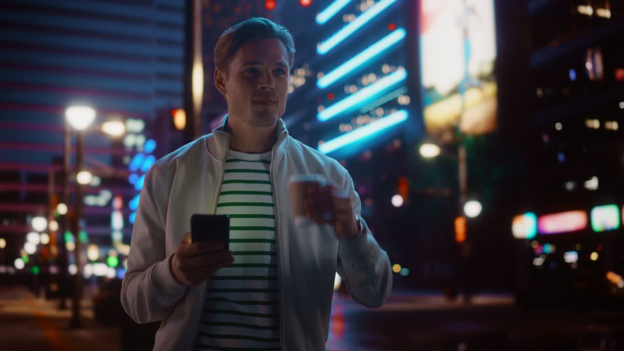 Portrait of Man Using Smartphone, Drinking Beverage, Walking Through Night City Street Full of Neon Light. Smiling Stylish Man Using Mobile Phone, Social Media, Online Shopping, Texting on Dating App