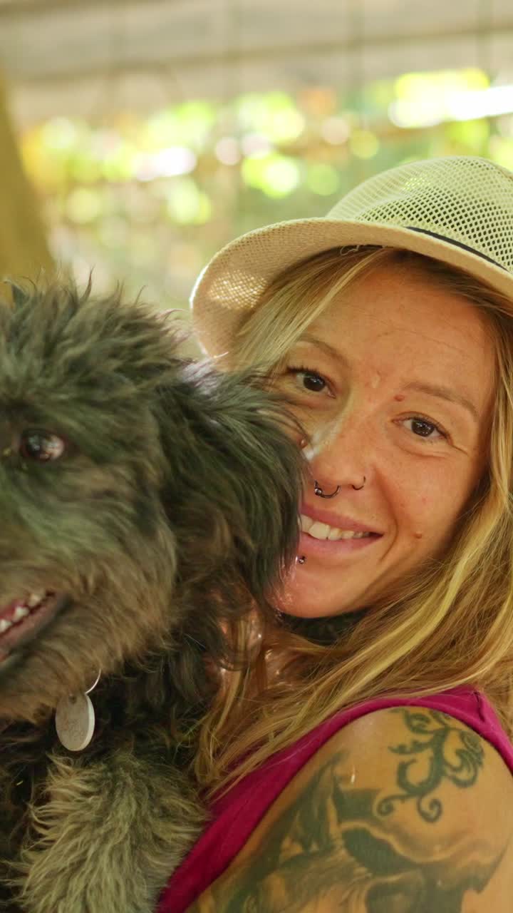 Donna felice con cappello e tatuaggi che tiene in braccio il suo cane all'aperto