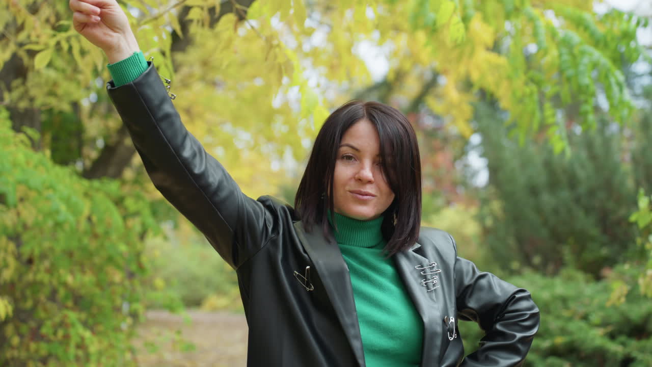Donna di colore sicura di sé con giacca di pelle che assume pose audaci in un parco autunnale con dolcevita verde, mani sui fianchi e braccio alzato, trecce che incorniciano il viso, sfondo di fogliame, spontanea