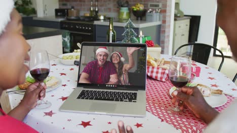 african american couple with wine using laptop for christmas video call with couple on screen