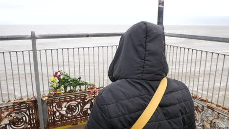 unrecognizable person in a hood contemplating a bouquet of flowers left in tribute. the gray sea and sky contribute to the solemn and reflective atmosphere of loss and bereavement after a death.