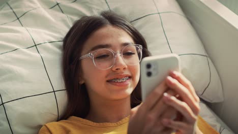 caucasian teenage girl browsing phone while lying on bed on her back and smiling