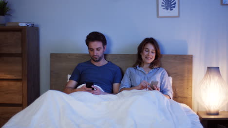 Pareja-Caucásica-Sentada-En-La-Cama-Debajo-De-Las-Mantas-Y-Usando-Teléfonos-Inteligentes-Por-La-Noche