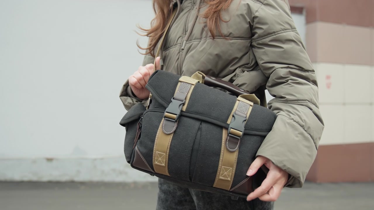 White woman adjusts black messenger bag while wearing olive puffer jacket, walking past concrete wall hands steady as she slides strap and fastens buckles, scenes suggest commuter, freelance