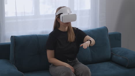 mujer con pantalla hmd en la cabeza está sentada en el sofá en el apartamento gesticulando con las manos tecnología moderna de realidad virtual